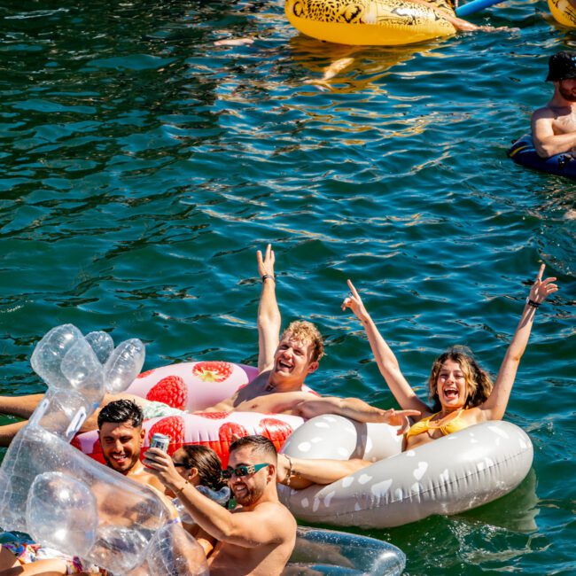 A group of people are enjoying a sunny day on the water, floating on various inflatable pool floats, including a panda, a strawberry, and an inflatable swan. They are smiling and laughing amidst others on similar floats. The clear blue water enhances the vibrant scene at Sydney Harbour Boat Hire The Yacht Social Club.