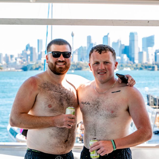 Two shirtless men with drinks pose on a boat with Sydney's skyline in the background. One man has sunglasses and a beard, while the other has short hair and a tattoo on his chest. They appear relaxed and are smiling at the camera during The Yacht Social Club event. The setting is sunny and bright.