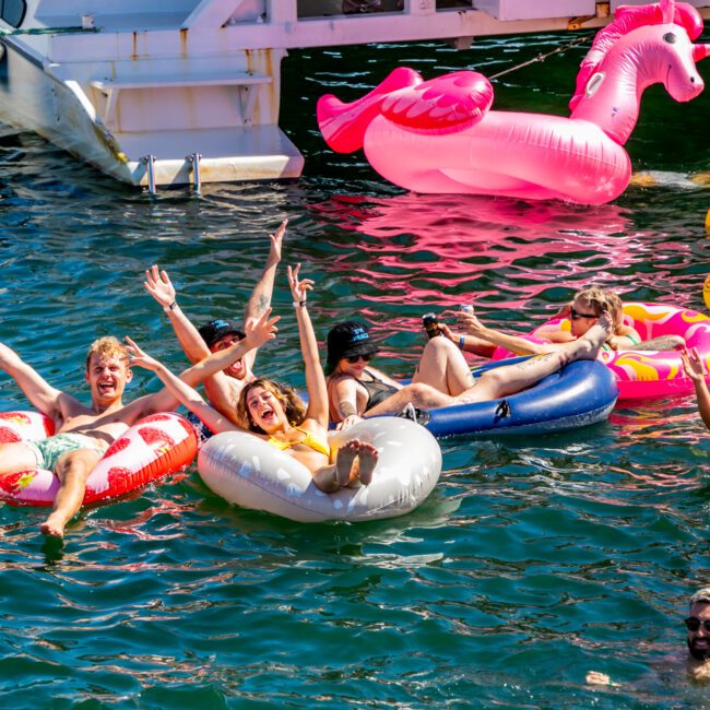 A group of people enjoying a sunny day on a lake, floating on colorful inflatables like a white swan and pink flamingo. Some are waving their hands in the air and laughing, while others relax on their floats. The Yacht Social Club’s luxurious atmosphere adds to the vibrant, clear water experience.