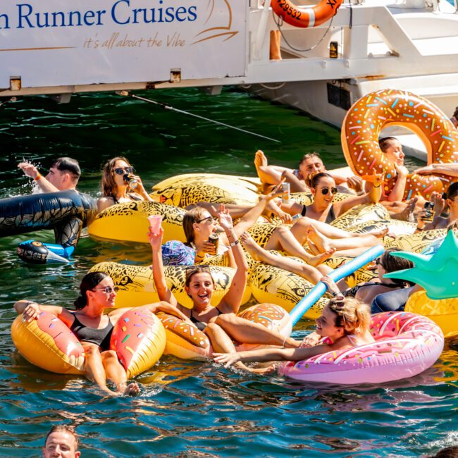 A group enjoys a sunny day, floating on inflatable pool toys near a boat. They are laughing, drinking, and socializing. The boat has a sign that reads "The Yacht Social Club Sydney Boat Hire." The scene is lively, colorful, and cheerful.