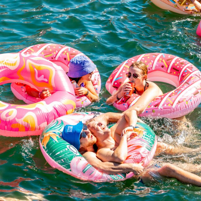 People are floating on inflatable rings in a body of water, enjoying a sunny day. One person is wearing a blue hat, another has sunglasses and a drink. The inflatables are colorful, with some resembling donuts. The mood is fun and relaxed, reminiscent of Boat Parties Sydney at The Yacht Social Club.