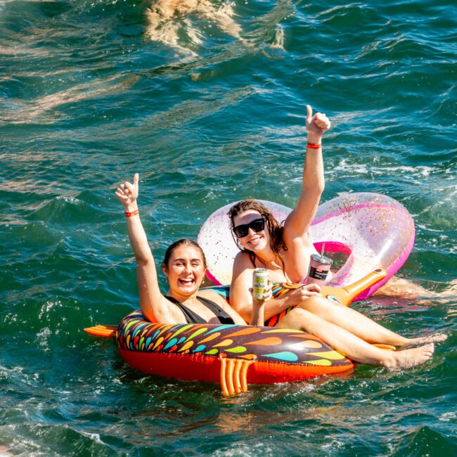 Two women float on inflatable pool tubes in a lake, smiling and holding up their arms joyfully. One tube is striped, the other heart-shaped. The water glistens under the sun with others enjoying it in the background—a perfect day reminiscent of a Luxury Yacht Rentals Sydney adventure.