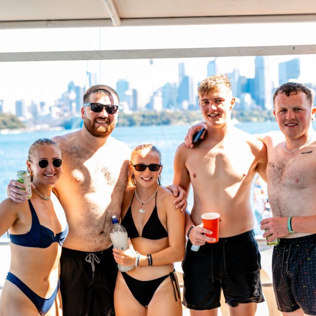 A group of five people in swimwear smile and pose together on a boat with a cityscape in the background. The weather appears sunny, and they are holding drinks, enjoying a day out on the water with Sydney Harbour Boat Hire The Yacht Social Club.