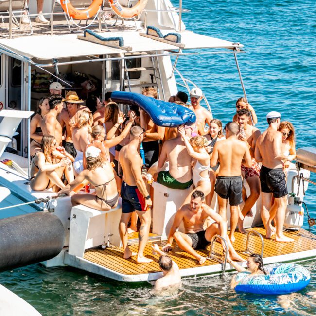 A lively group of people enjoy a sunny day on a yacht, gathered on its deck and swim platform. Some are in swimsuits, holding inflatable toys, and playing in the water. The clear blue sea surrounds the boat from The Yacht Social Club's luxury yacht rentals in Sydney, with a few people floating on inflatables nearby.