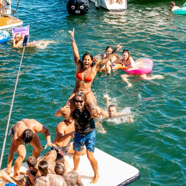 A lively group of people enjoys a sunny day on the water. Some are on a floating pad, with a man giving a woman a shoulder ride as she raises her hands in excitement. Others swim or float in inner tubes, with boats from Luxury Yacht Rentals Sydney in the background. Everyone is in swimwear.