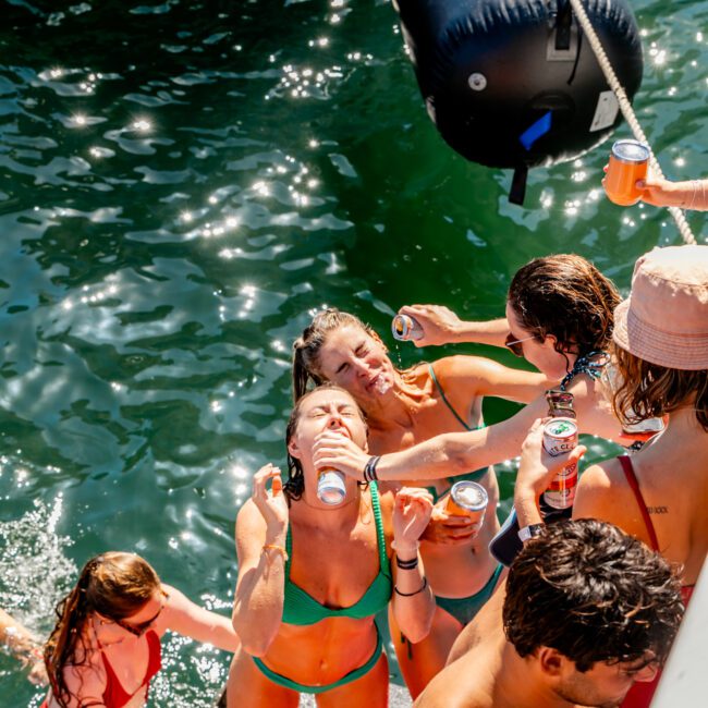 A group of people in swimsuits are enjoying a sunny day on the water. Some are in the water, while others are on a boat rented from The Yacht Social Club Sydney Boat Hire, holding drinks and smiling. One person is pouring a drink into another's mouth. The scene is lively and relaxed, with a festive atmosphere.