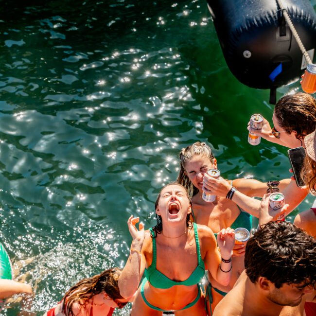 A group of people in swimwear enjoying a sunny day on a boat. One woman in the center is laughing and gesturing with her hands while others around her are holding drinks and smiling. The boat is surrounded by green water, with some people swimming near the boat. It's a perfect outing with Boat Parties Sydney The Yacht Social Club.