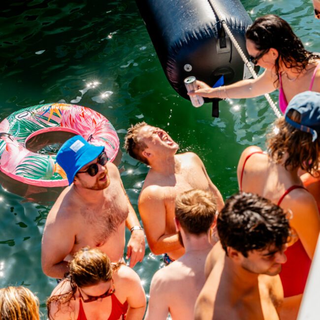 A group of friends in swimwear is gathered on a boat, enjoying the sunny weather. One person is handing a drink to another. An inflatable float with a colorful pattern is seen in the water as part of The Yacht Social Club's Boat Parties Sydney. Everyone appears to be having a good time.