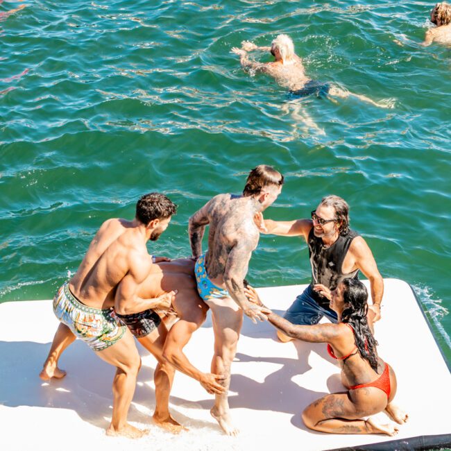 A group of people are on a floating platform in the water. One person is being pulled up by two others, while a woman in a red bikini and another person look on. Several individuals are swimming in the surrounding greenish-blue water under sunny conditions, enjoying The Yacht Social Club Event Boat Charters.