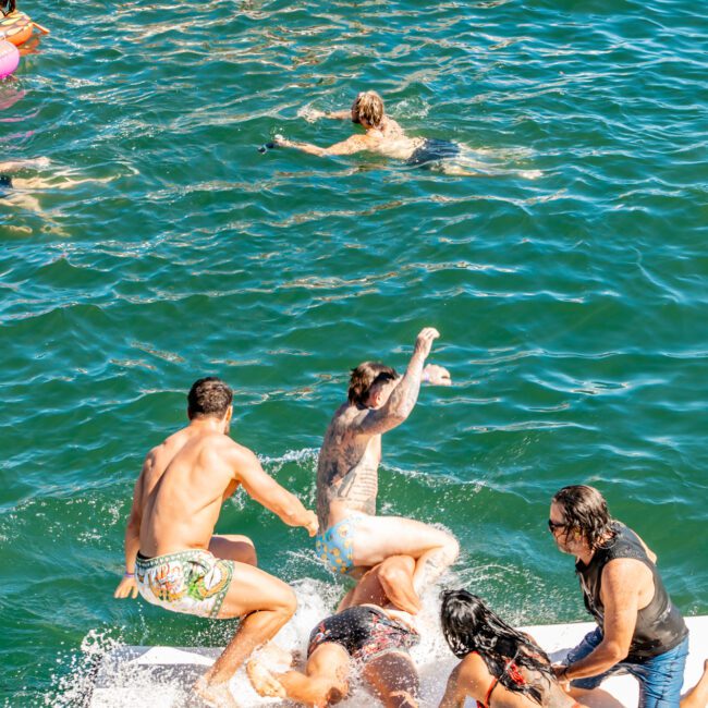 A group of people is playing and splashing in the clear blue water near boats. Some are on a floating platform, with two men splashing another man who is falling. A woman in a red swimsuit and a man in a black shirt are helping. Others swim in the background, enjoying Boat Parties Sydney The Yacht Social Club.