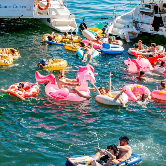 A lively scene on the water with numerous people enjoying a sunny day on inflatable floats, primarily in the shapes of pink flamingos and unicorns. Nearby, two white boats from The Yacht Social Club Sydney are filled with more people lounging. The water sparkles in a clear, vibrant blue.