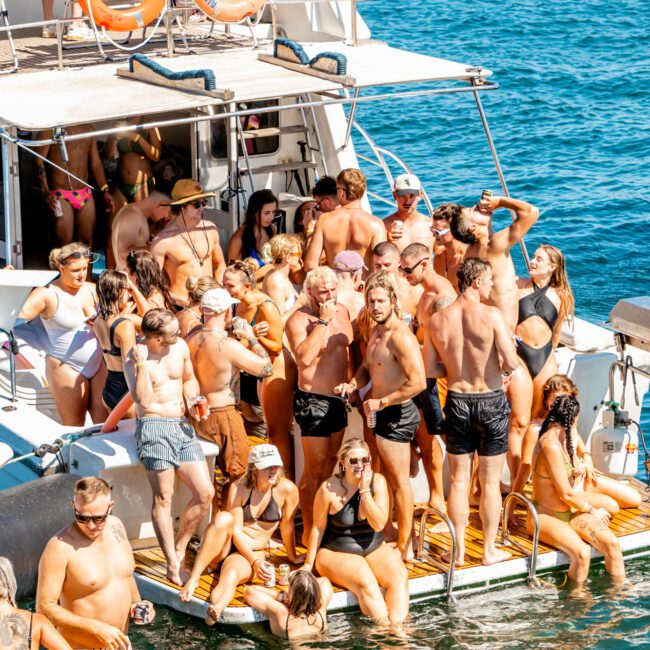 A large group of people in swimwear are on a boat and the back dock, gathered closely together. They appear to be enjoying a sunny day on the water. Some are lounging or sitting while others stand and chat. Floatation devices are visible nearby at The Yacht Social Club Event Boat Charters.