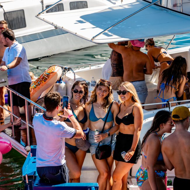 A group of people enjoys a sunny day on a boat. Three women in swimsuits pose for a photo taken by a man. Other party-goers with The Yacht Social Club Sydney Boat Hire are on nearby boats and in the water, some floating on inflatable rafts. The atmosphere is lively and festive.