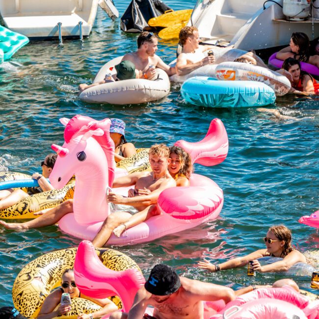 A lively group of people enjoy a sunny day on the water, lounging on inflatable pool floats, including an eye-catching pink unicorn. They are smiling, chatting, and holding drinks near boats from The Yacht Social Club Event Boat Charters, with others in colorful floats and swim rings surrounding them.