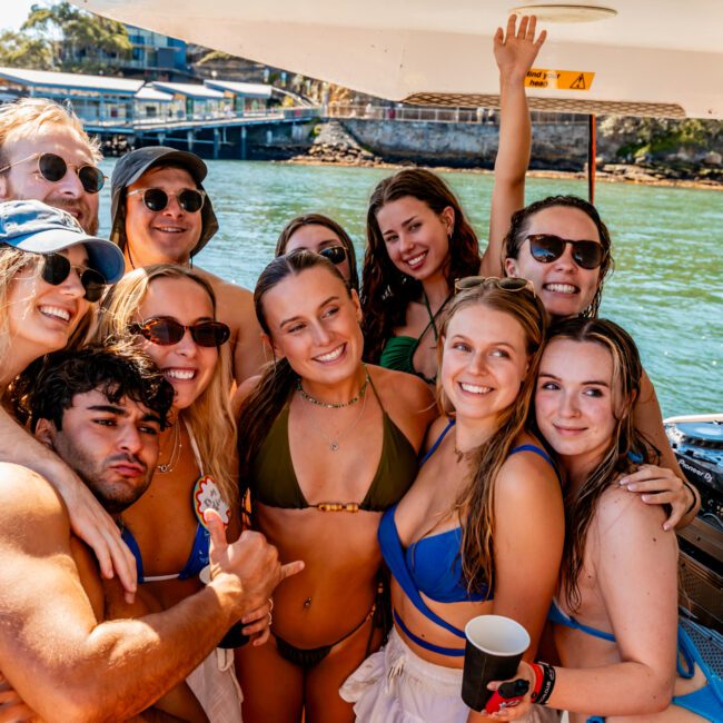 A group of people smiling and posing for a photo on a boat from The Yacht Social Club Sydney Boat Hire. Some are wearing swimsuits and sunglasses, while others have on hats. The background features water, another boat, and a shoreline with buildings. One person appears to be DJing in the background.