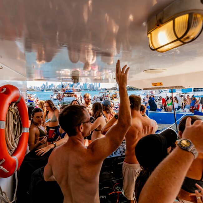 A lively group of people is dancing and socializing on The Yacht Social Club Event Boat Charters, enjoying the sunny weather. Some are wearing swimsuits, and the background shows another boat and a city skyline. A life preserver is mounted on the boat's wall, and the mood is festive.