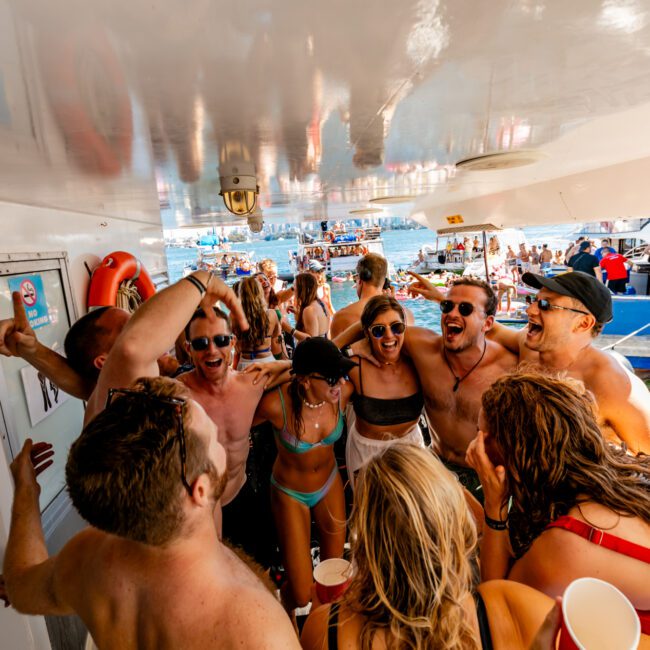 A lively group of people in swimsuits and summer attire are gathered closely together, smiling and enjoying a party on a boat. Some are holding drinks, with The Yacht Social Club Event Boat Charters providing the festive atmosphere against the backdrop of blue water.