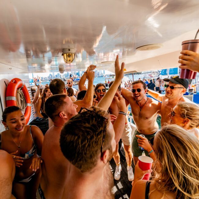 A lively group of young adults is on a boat, dancing and celebrating. The atmosphere is energetic, with drinks in hand and smiles all around. The sun shines brightly, reflecting off the water in the background, while life vests hang on the wall. Experience it with The Yacht Social Club Sydney Boat Hire for luxury yacht rentals.