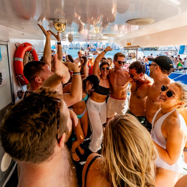 A group of people in swimwear are dancing and celebrating on a boat under a sunny sky. They are closely gathered, some raising their hands, with the city skyline visible in the background. Smiling faces and a festive atmosphere dominate the scene at The Yacht Social Club event, with a life preserver mounted on a nearby wall.