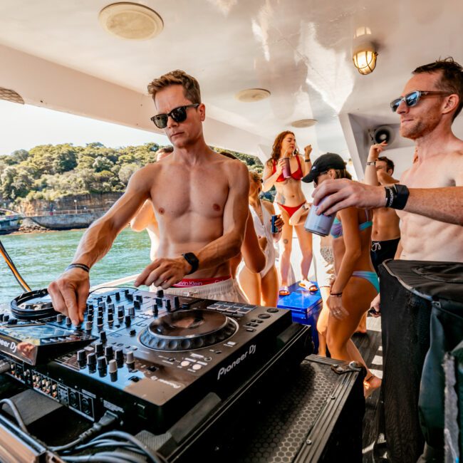 A shirtless man is DJing on a boat at The Yacht Social Club Event Boat Charters, with another shirtless man holding a drink nearby. Several people in swimwear are dancing and enjoying the sunny day on the water, with scenic greenery and buildings visible in the background.