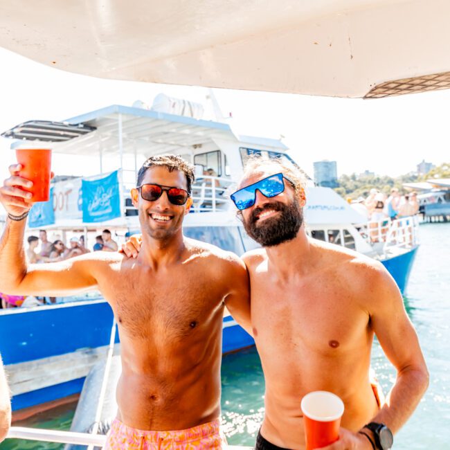 Two smiling shirtless men wearing sunglasses and holding red cups, standing arm-in-arm at a Yacht Social Club event. They pose in front of a blue and white boat with people onboard. The background reveals clear skies and calm water, indicating a sunny day on the water.