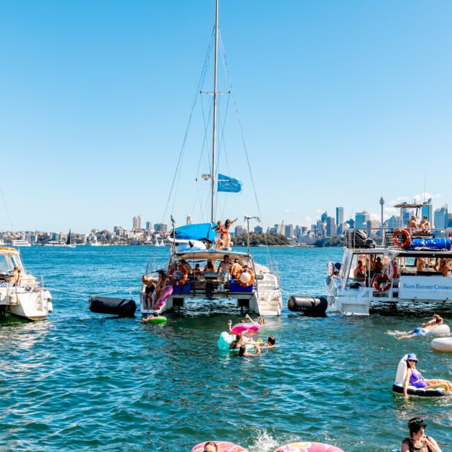 People enjoy a sunny day on the water, swimming and lounging on floats near three anchored boats. The boats, part of Boat Parties Sydney The Yacht Social Club, are decorated with blue flags and colorful balloons. A city skyline is visible in the background under a clear blue sky.