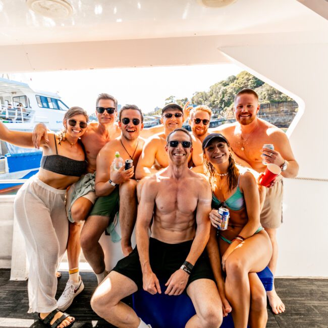 A group of nine people, wearing swimsuits and casual summer attire, pose cheerfully on a luxury yacht from The Yacht Social Club Sydney Boat Hire. Some are holding drinks, and all are smiling at the camera. The background features another boat, lush greenery, and clear blue skies, suggesting a fun day out on the water.