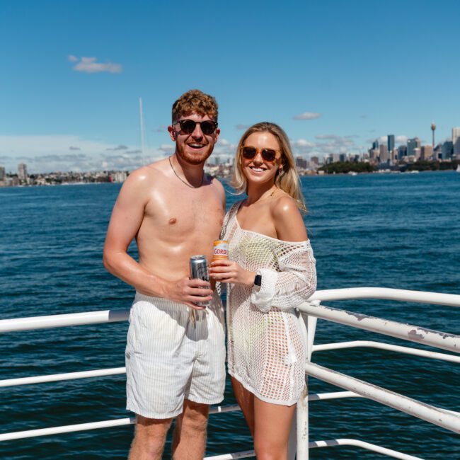 A man and woman, both wearing sunglasses, smile and hold drinks while standing on a boat with a body of water and city skyline in the background. The man is shirtless and in shorts, while the woman is wearing a swimsuit and a white cover-up. It appears to be a sunny day, perfect for The Yacht Social Club Sydney Boat Hire.