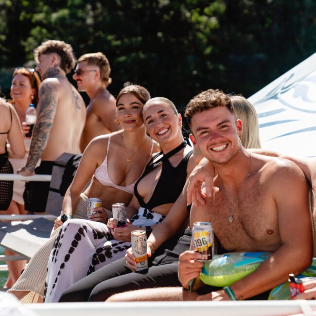 A group of people in swimwear are smiling and holding canned drinks while sitting on a boat under the sun. Trees are visible in the background. Everyone looks happy and relaxed, enjoying The Yacht Social Club Event Boat Charters outdoor setting.