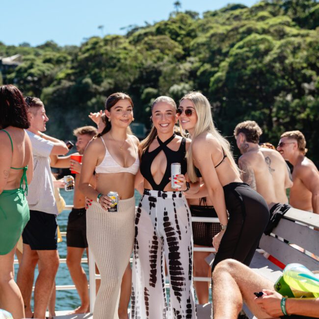 Three women are standing together on a boat, smiling at the camera and holding drinks. Dressed in summer attire, they are surrounded by others enjoying a sunny day on the water. The background features lush green trees along the shoreline, capturing the essence of The Yacht Social Club Event Boat Charters.