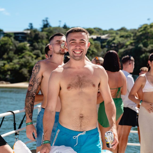 A shirtless man in blue swim trunks stands on a boat, smiling and holding a drink can. Other casually dressed individuals are around him, some in swimwear. The background features water and a tree-covered shoreline under a clear blue sky, perfect for Luxury Yacht Rentals Sydney's spectacular escapes.