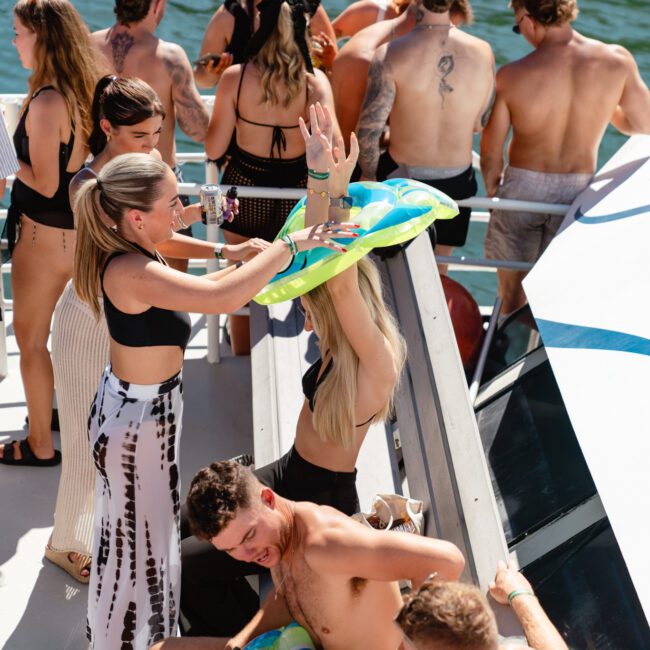 A group of people enjoying a boat party hosted by The Yacht Social Club Sydney Boat Hire. Some are wearing swimsuits and summer attire. One person is playfully wearing an inflatable ring on his arms, while others are socializing and laughing. The background shows water and part of the boat decorated with blue patterns.