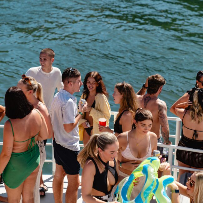 A group of people are enjoying a sunny day on a boat from Sydney Harbour Boat Hire The Yacht Social Club. They are dressed in swimsuits, swim trunks, and casual summer clothes, holding drinks, and chatting. The boat is on a body of water, with sunshine reflecting off the surface.