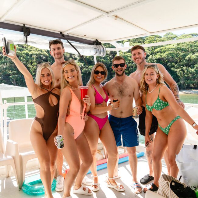 A group of seven people, dressed in swimwear, stand together on a boat under a canopy. They are smiling and holding drinks, with lush green trees and water visible in the background. Experience this with The Yacht Social Club Sydney Boat Hire for an unforgettable time on the water.