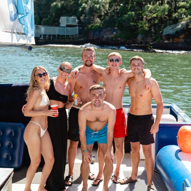 A group of six people, four men and two women, in swimwear are posing and smiling on a boat from The Yacht Social Club with a scenic forested shoreline in the background. Some hold drinks, and a few floatation devices are visible on the boat. It is a sunny day.