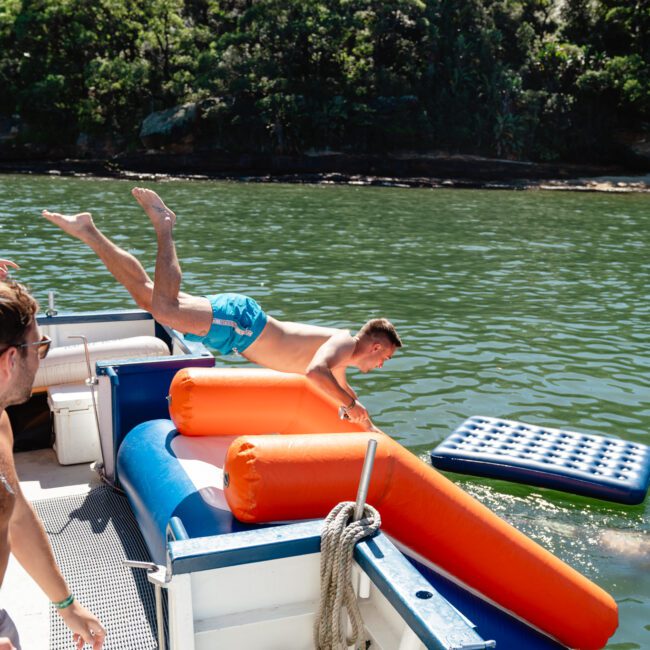 A man in blue swimming trunks is mid-dive off a luxury yacht into a lake with lush green trees in the background. He is diving over an inflated orange structure on the boat, while another person watches from the side. Various floatation devices are in the water, enjoying The Yacht Social Club Sydney Boat Hire.