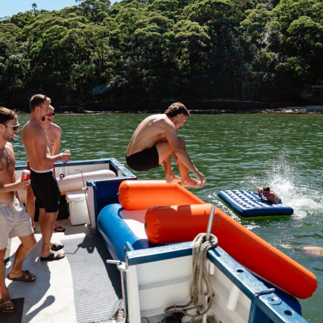 A group of young men are having fun on a luxury yacht on a sunny day. One person is jumping into the water, where an inflatable mattress is floating. The background shows a lush green forest lining the shore. The mood is playful and energetic.