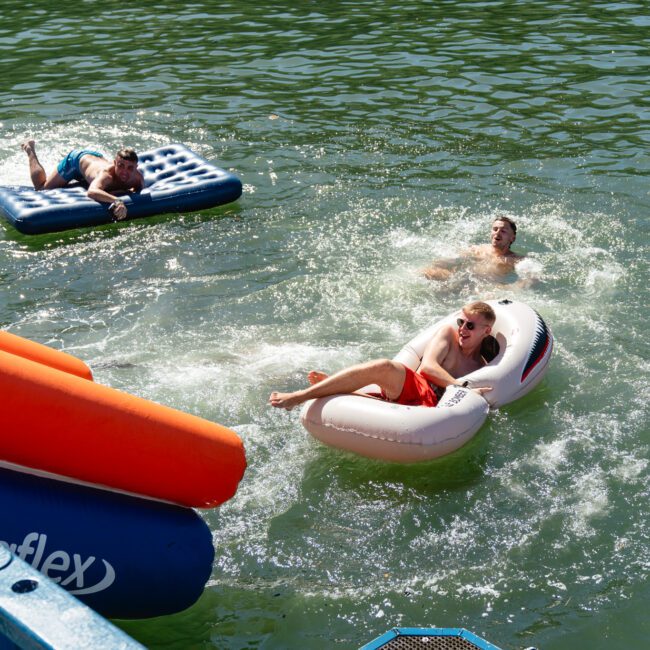 Three people are enjoying a sunny day in the water. One person is lying on a blue inflatable mattress, another is relaxing on a white inflatable float with a shark design, and the third person is swimming nearby. The scene is energetic and joyful, perfect for The Yacht Social Club Event Boat Charters.