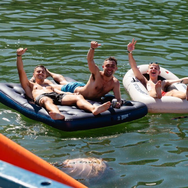 Three people are lying on inflatable mattresses in a calm, greenish lake, smiling and raising their arms in the air. Bright colored objects float nearby and a corner of a life raft is visible. They appear to be enjoying a sunny day reminiscent of Boat Parties Sydney The Yacht Social Club.