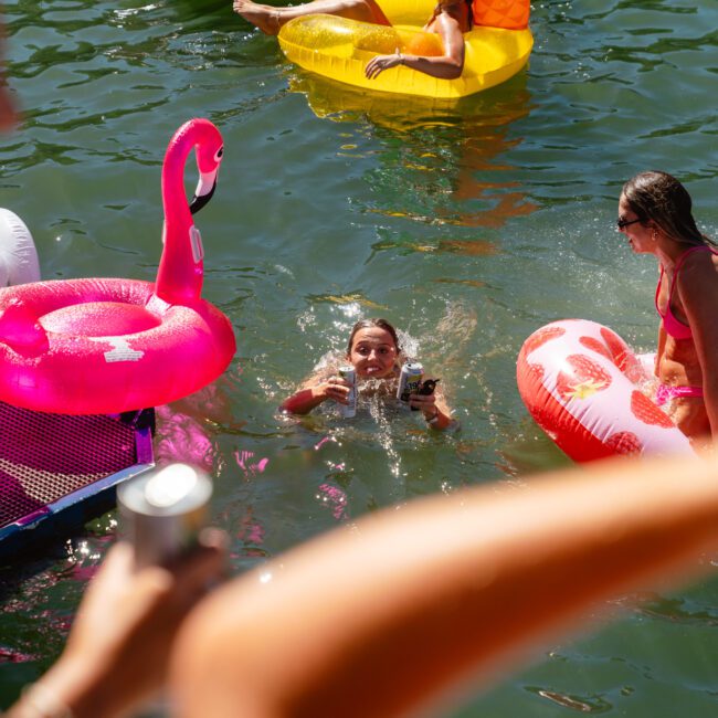 A group of people enjoy a sunny day by the water, surrounded by colorful pool floats. A person in the water wearing goggles holds up a phone and waves, while another lounges on a heart-shaped float. The scene is festive and cheerful, reminiscent of adventures with Sydney Harbour Boat Hire The Yacht Social Club.