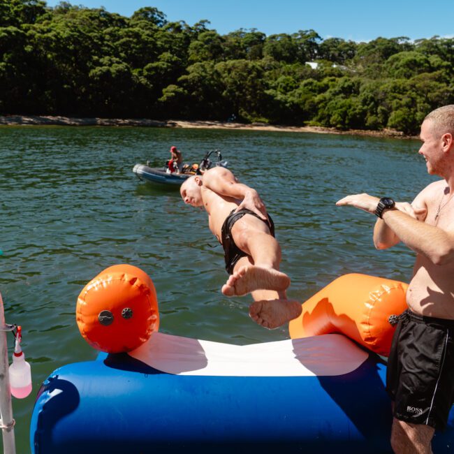 Two men are on a floating water slide by a lake surrounded by a forest. One slides down into the water, while the other stands at the top, gesturing with his hands. A yellow inflatable float can be seen in the water, and a small boat is in the background, reminiscent of Boat Parties Sydney The Yacht Social Club.