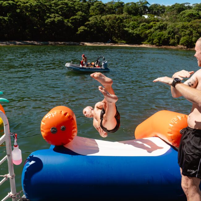 A person jumps off a floating platform while performing a backflip. Another person stands nearby, watching and gesturing mid-action. In the background, a boat from The Yacht Social Club with a few people and an inflatable float are seen on the water near a forested shoreline.