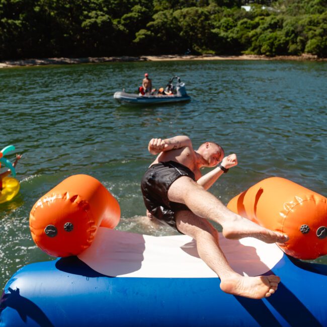 A man slides off an inflatable water float into the lake, surrounded by lush greenery. Another person in an inflatable tube with palm tree decorations is in the water, while a group on a small boat is in the background. It's a sunny day filled with fun at The Yacht Social Club Sydney Boat Hire.