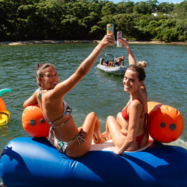 Two women in swimwear sit on an inflatable float in a lake, holding up drinks and smiling at the camera. Surrounding them are people swimming and relaxing on various inflatables. In the background, a boat from The Yacht Social Club Event Boat Charters is visible against a green, forested shoreline.