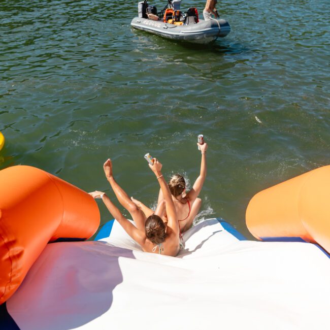 Two people slide down an inflatable structure into a lake, each holding a can. Nearby, a group floats on an inflatable palm tree and someone paddles in an inflatable boat. The scene is sunny and festive, reminiscent of the lively vibes at The Yacht Social Club Sydney Boat Hire events.