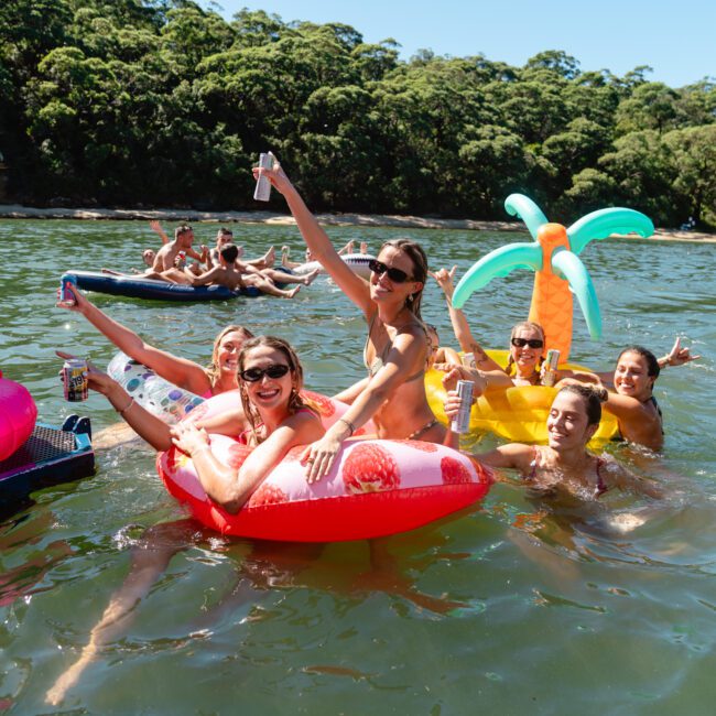 A group of smiling people in swimsuits enjoy a sunny day in the water. Some are floating on inflatable pool floats, including a shrimp and a palm tree. Holding drinks and having fun surrounded by greenery, it feels like The Yacht Social Club Event Boat Charters or Luxury Yacht Rentals Sydney experience.