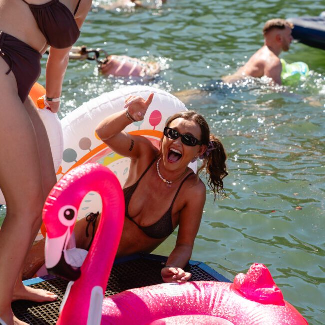 Two women in swimsuits enjoy a sunny day on the water, holding vibrant pool floats, including a pink flamingo-shaped one. They smile and gesture enthusiastically amidst a festive atmosphere. Others swim and relax in the background, showcasing fun outdoor activities enjoyed at Sydney Harbour Boat Hire The Yacht Social Club.