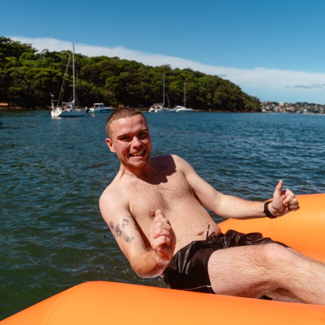 A smiling shirtless man with short hair and tattoos on his upper arms gives two thumbs up while sitting on an orange inflatable boat in a sunny bay. In the background, sailboats and a Luxury Yacht Rentals Sydney vessel are anchored near a forested shoreline. The sky is clear and blue.