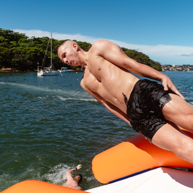 A shirtless man in black shorts is captured mid-air above the water, appearing to jump or fall from an orange and white inflatable object during a Sydney Harbour Boat Hire The Yacht Social Club event. In the background, there’s a scenic view of a bay with boats, trees, and a clear blue sky.