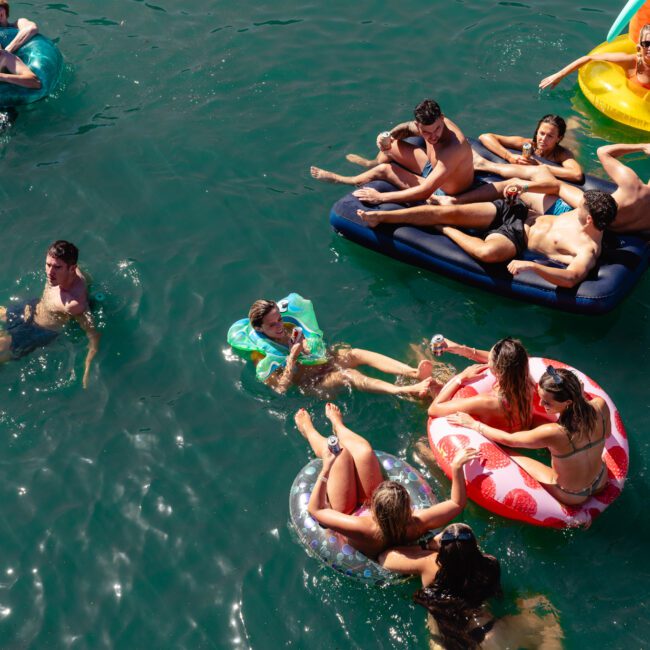 Aerial view of a group of people in a body of water, floating on different inflatable items, such as rings and loungers. They are enjoying a sunny day, relaxing and interacting with each other at The Yacht Social Club Event Boat Charters. The water appears to be calm and clear.